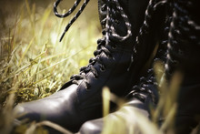 Black Rough Leather Army High Boots With Black And White Laces Standing In The Grass, Illuminated By Sunlight.