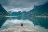 Fototapeta  - Kobieta samotnie siedzi na drewnianym pomoście nad górskim jeziorem Traunsee w Austrii w pochmurny dzień	