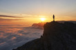 © kravtzov - silhouette of hiker woman with trekking poles stands at rock and looks at aerial view in the mountains above the clouds. Mountains sunset landscape with tourist silhouette in orange sun light.