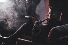 Groomed Bearded Man Is Relaxing On Lounge Near Fireplace While Smoking Hookah. He Has Tattoo On His Hand.