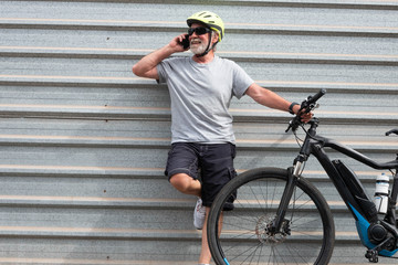 Adult senior caucasian man enjoying e bike bicycle in the city. Resting and talking on mobile phone. White hair and beard. Yellow helmet. Gray background in the street