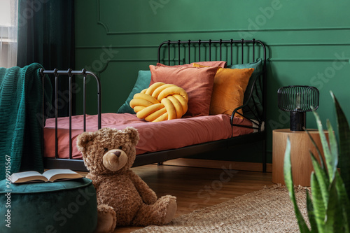 Fotografía Big, brown teddy bear and an open book on a green, velvet pouf in front of a metal frame bed in child's bedroom interior