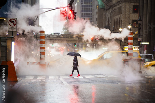 A woman with an umbrella and red high heels shoes is crossing the 42nd street in Manhattan. Cars and steam coming out from from the manholes in the background. New York City, Usa.