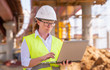 © denklim - Girl foreman with laptop at construction site. female engineer works on computer under overpass under construction.