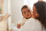 Smiling loving young African mom with curly hair standing in room and holding adorable baby boy with chubby cheeks