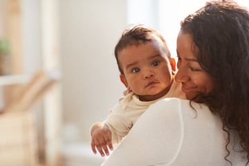 smiling loving young african mom with curly hair standing in room and holding adorable baby boy with