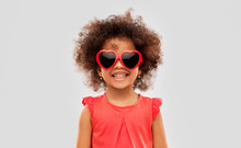 Childhood, Valentine's Day And Summer Concept - Happy Little African American Girl In Heart Shaped Sunglasses Over Grey Background