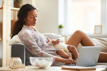 Content Pensive Busy Young Black Mother In Cotton Homewear Sitting On Sofa And Working With Laptop While Baby Sleeping