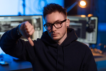 Poster - cybercrime, hacking and technology concept - young asian hacker wearing glasses and hoodie in dark room with computers showing thumbs down at night
