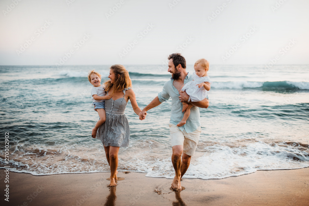 Young family with two toddler children walking on beach on summer holiday. - obrazy, fototapety, plakaty 