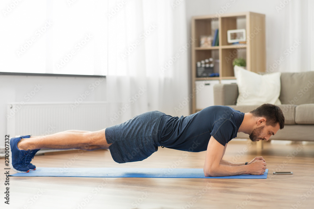 sport, fitness and healthy lifestyle concept - man looking at smartphone and doing plank exercise at home - obrazy, fototapety, plakaty 