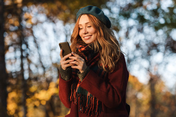 Wall Mural - Happy young pretty woman walking outdoors in autumn spring park using mobile phone chatting.