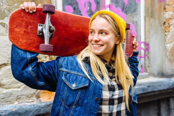 Nice cute lovely attractive magnificent blond woman in yellow hat blue denim jacket with red longboard posing against aged vintage abandoned building facade with pink inscription