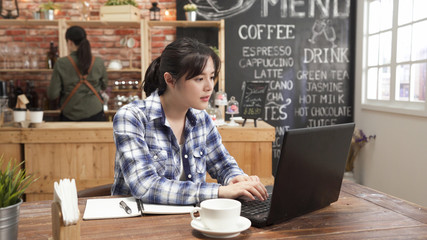 Image of happy woman using laptop while sitting at cafe bar. Young asian girl sitting in coffee shop and working on computer notebook. elegant barista in apron back view busy making beverage counter