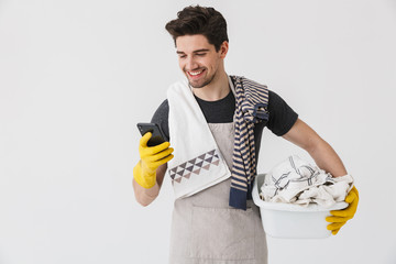 Wall Mural - Photo of smiling young man wearing yellow rubber gloves using smartphone while carrying laundry basket with clothes