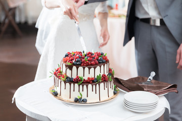 Wall Mural - Bride and groom cut beautiful rustic wedding cake on wedding banquet. The cake is decorated with fresh berry, strawberry, raspberry and chocolate. Fashionable luxury stylish cake