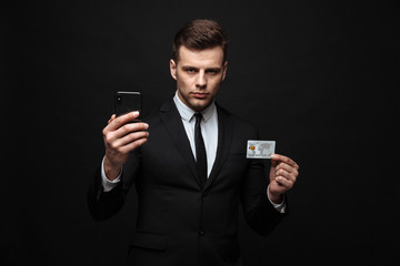 Poster - Portrait of serious young businessman dressed in formal suit holding cellphone and credit card isolated