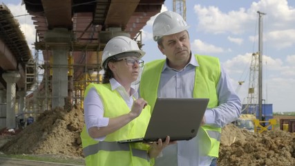 Wall Mural - Closeup of man and woman working on background of construction overpass. Builders discussing future work plan for erection of bridge using computer.