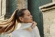 Fashionable woman with long hair in ponytail hairstyle wearing trendy hoop earrings, white turtleneck sweater, posing in street of European city. Copy, empty space for text
