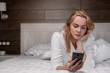 © spaskov - An attractive middle-aged blonde woman in a white robe is lying on a bed in a bedroom and using a smartphone. Smiles and laughs.