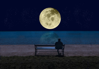 man sitting on a bench by the sea in the night and looking at a big shining full moon with stars ref