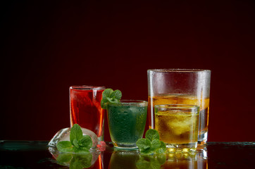 Three different alcoholic drinks in clear glasses with ice and mint on red background