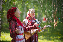 Two Young Woman In Traditional Russian Clothes Singing In The Forest. One Of Them Playing Balalaika