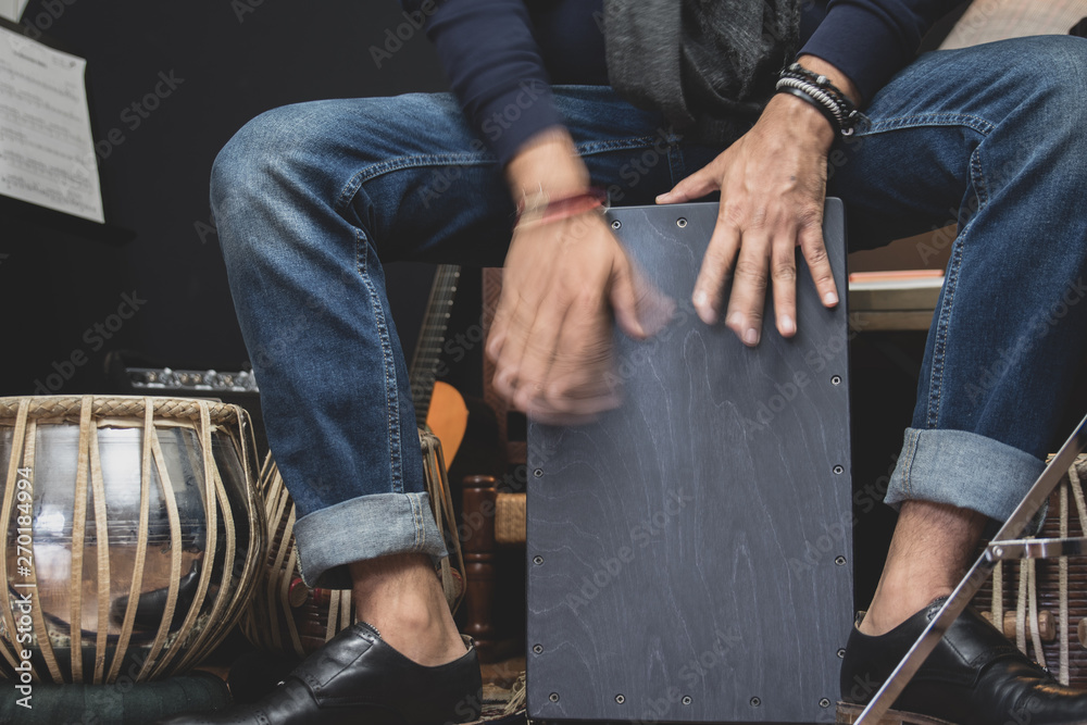A stylish musician in denim and double monk shoes plays the Cajon, a Peruvian drum used commonly with Spain’s Flamenco dance.  - obrazy, fototapety, plakaty 