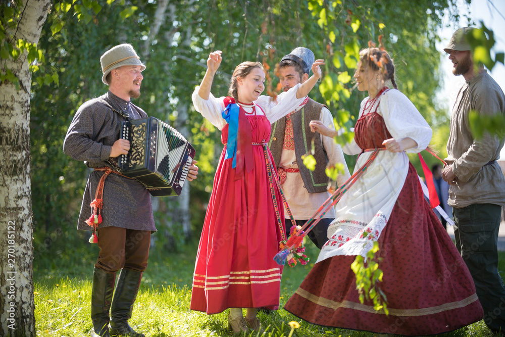 People in traditional Russian clothes are dancing in the woods - one of them plays the accordion and singing - obrazy, fototapety, plakaty 