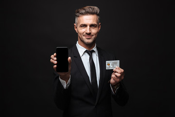 Business man posing isolated over black wall background using mobile phone holding credit card.