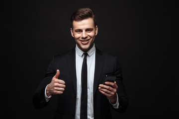 Poster - Handsome business man posing isolated over black wall background using mobile phone showing thumbs up.