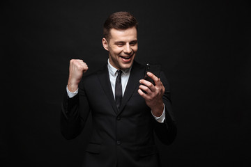 Wall Mural - Handsome business man posing isolated over black wall background using mobile phone showing winner gesture.