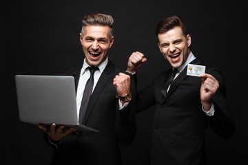 Poster - Handsome excited happy two business men posing isolated over black wall background using laptop computer holding credit card.