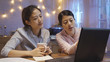 © PRPicturesProduction - Two young asian women sitting at wooden kitchen table talking discussing at night. smart girl in pink pajama show point partner friend information on laptop screen. Teamwork freelancers working.
