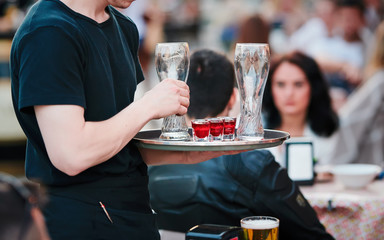 Waiter with tray takes empty beer glasses and brings cocktails to the guests of restaurant on the summer terrace. Service in cafes and restaurants. Man holding the tray with red cocktails.