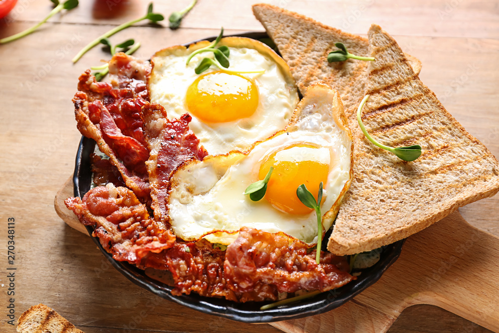 Frying pan with tasty eggs, bacon and toasts on table