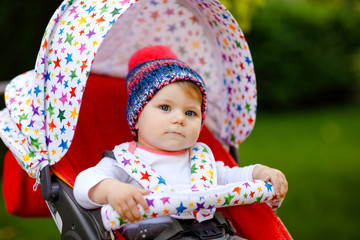 Canvas Print - Cute healthy little beautiful baby girl with blue warm hat sitting in the pram or stroller and waiting for mom. Happy smiling child with blue eyes. baby daughter going for a walk with family