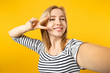 © ViDi Studio - Close up selfie shot of cheerful young woman in striped clothes showing victory sign isolated on yellow orange wall background in studio. People sincere emotions lifestyle concept. Mock up copy space.