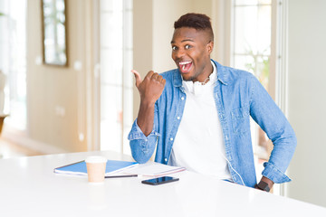 Sticker - African american student man studying using notebooks and wearing headphones smiling with happy face looking and pointing to the side with thumb up.