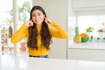 Sticker - Young beautiful woman at home on white table covering ears with fingers with annoyed expression for the noise of loud music. Deaf concept.