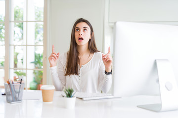 Poster - Beautiful young woman working using computer amazed and surprised looking up and pointing with fingers and raised arms.