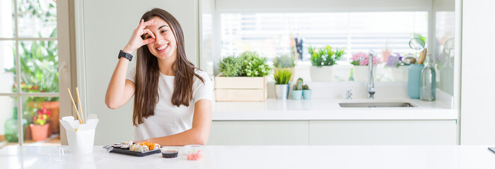 Canvas Print - Wide angle picture of beautiful young woman eating asian sushi from delivery doing ok gesture with hand smiling, eye looking through fingers with happy face.