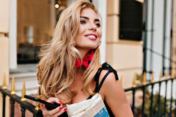 Close-up portrait of spectacular woman with blonde shiny hair looking up and laughing near restaurant. Cheerful lady with blue eyes holding phone and documents, walking in square.