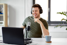 technology, mass media and podcast concept - happy young male audio blogger in headphones with laptop computer and microphone broadcasting at home office