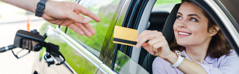 panoramic shot of cheerful woman sitting in car and giving credit card to worker at gas station