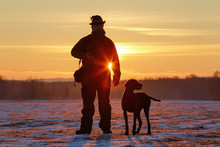 A Hunter In The Hat With A Gun And His German Shorthaired Pointer Dog Breed Friend. Beautiful Silhouettes On The Background Of The Dawn. Amazing Winter Morning.