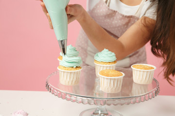 Wall Mural - Close up cropped housewife female chef cook confectioner or baker in apron white t-shirt decorating cake cupcake at table isolated on pink pastel background in studio. Mock up copy space food concept.