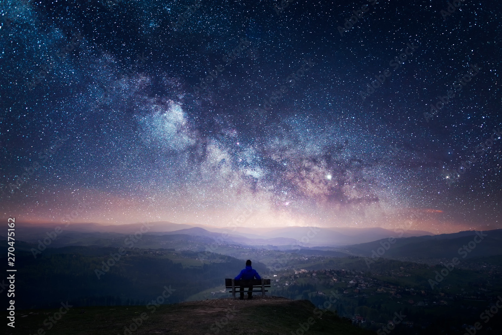 A man sitting on a bench staring at a starry sky with a Milky Way and a mountain landscape - obrazy, fototapety, plakaty 
