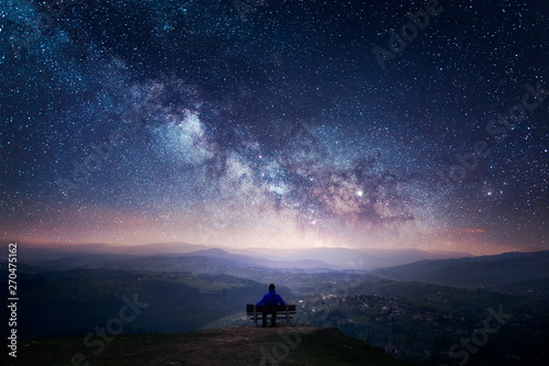 Cuadros en Lienzo A man sitting on a bench staring at a starry sky with a Milky Way and a mountain