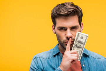 handsome man doing silence gesture and holding dollar banknotes Isolated On yellow